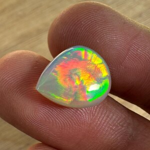 May include: A pear-shaped opal gemstone with a translucent, milky white base. The stone displays vibrant internal colors of red, orange, yellow, and green, creating a play-of-color effect. The opal is held between two fingers.