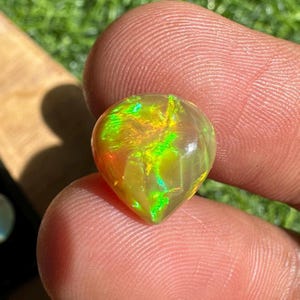 May include: A pear-shaped, faceted opal gemstone. The gem displays a vibrant play-of-color with flashes of green, yellow, and orange hues. The opal is held between two fingers, showcasing its polished surface and internal iridescence. The gemstone is set against a blurred background.
