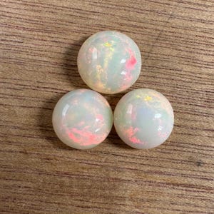 May include: Three round opals with a pearly white base color and flashes of pink, yellow, and green. The opals are set against a wooden surface, showcasing their iridescent qualities. These gemstones are ideal for jewelry making.