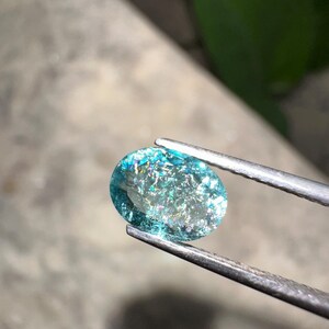 May include: An oval-shaped, iridescent gemstone held by tweezers. The gem has a light blue hue with sparkling inclusions, reflecting multiple colors. The tweezers are silver-colored, and the background is blurred.