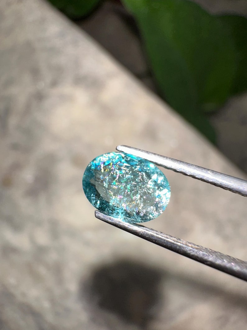May include: An oval-shaped, iridescent gemstone held by tweezers. The gem has a light blue hue with sparkling inclusions, reflecting multiple colors. The tweezers are silver-colored, and the background is blurred.