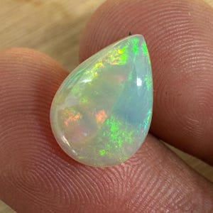 May include: A pear-shaped opal gemstone with a translucent, milky white base. The stone displays iridescent flashes of green, orange, and yellow. The opal is held between two fingers, showcasing its polished surface and natural beauty.