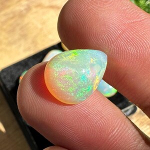 May include: A polished opal gemstone, displaying a spectrum of colors including orange, green, and yellow. The opal is held between fingers, showcasing its iridescent play-of-color. The gemstone has a smooth, rounded shape.