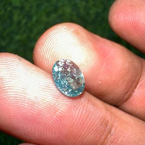 May include: An oval-shaped, translucent gemstone with a light blue hue and internal inclusions. The gem is held between fingers, showcasing its polished surface and natural imperfections. The gemstone is a beautiful example of natural mineral beauty.