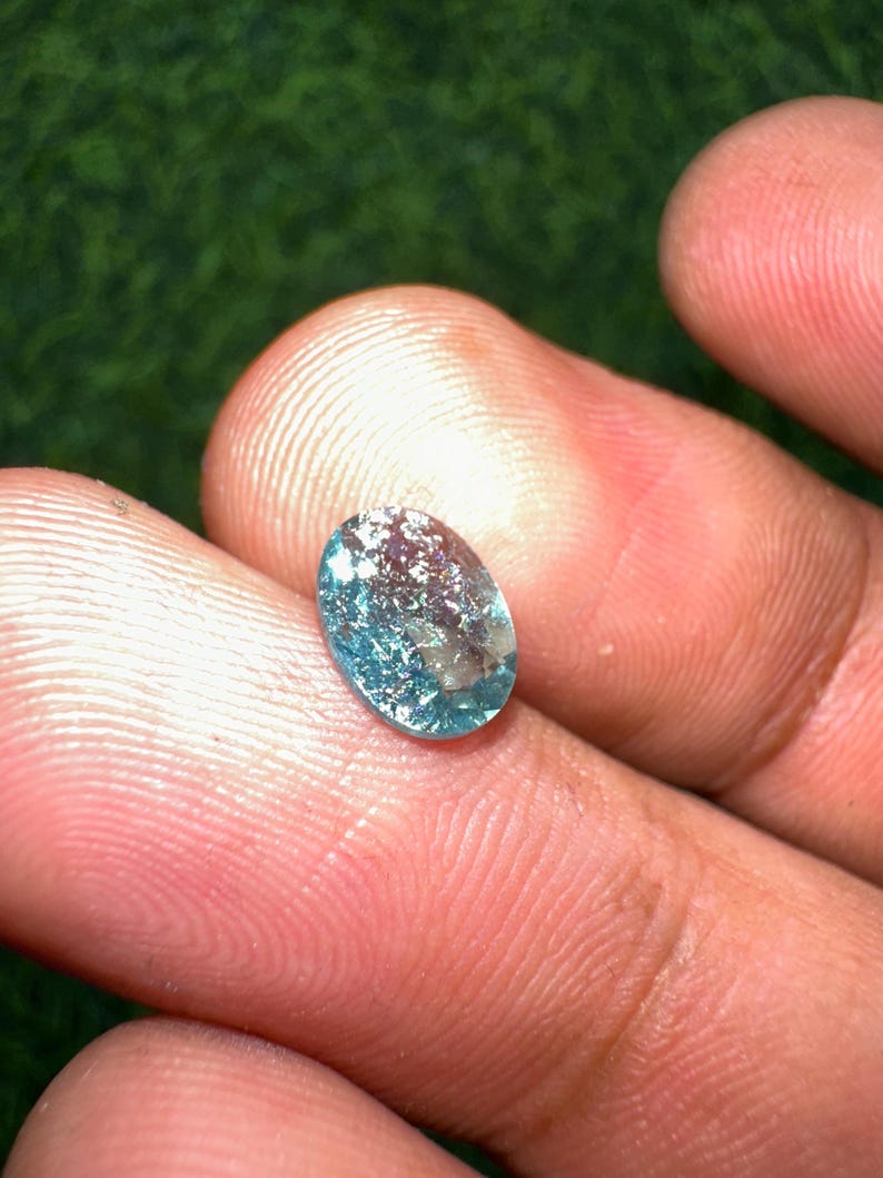 May include: An oval-shaped, translucent gemstone with a light blue hue and internal inclusions. The gem is held between fingers, showcasing its polished surface and natural imperfections. The gemstone is a beautiful example of natural mineral beauty.