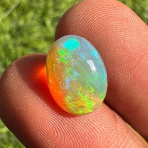 May include: An oval-shaped opal gemstone with a vibrant display of colors. The opal showcases a mix of orange, yellow, green, and blue hues, creating a shimmering effect. The gemstone is held between fingers, highlighting its size and shape.