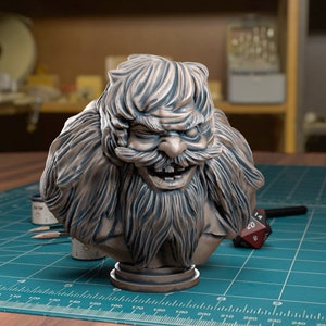 Könnte beinhalten: Detaillierte Büste eines bärtigen, grinsenden Fantasy-Charakters mit großem Schnurrbart und gerunzelter Stirn. Die Skulptur hat eine gedämpfte Bronzefarbe und steht auf einem runden Sockel. Die Büste steht auf einer grünen Schneidematte mit einem rot-schwarzen Würfel.