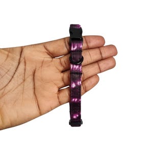 May include: A purple and white patterned pet collar with a black plastic buckle and a black metal D-ring. The collar is held in a hand against a white background.