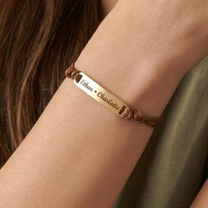 May include: A gold-colored bracelet with the names "Ethan • Charlotte" engraved on a rectangular plate. The plate is attached to a brown cord bracelet. The person is wearing a green shirt.