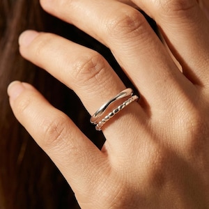May include: Two silver rings are displayed on a finger. The top ring has a smooth, wavy design, while the bottom ring features a textured, twisted pattern. The rings are worn on a hand, with the background slightly blurred.