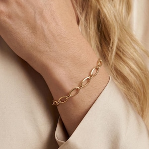 May include: A gold-coloured link bracelet. The bracelet features oval-shaped links. The bracelet is worn on a wrist. The background is a light beige colour.