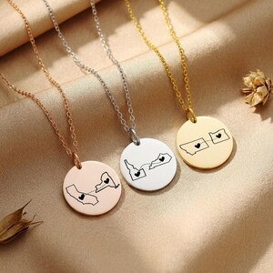 May include: Three circular pendant necklaces in rose gold, silver, and gold tones. Each pendant features two engraved state outlines connected by a dashed line, with a small black heart inside each state. The necklaces have delicate link chains.