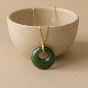 May include: A dark green circular jade pendant suspended from a thin gold-toned metal chain. The smooth, polished stone ring features a central hole and is attached to the chain by a small gold-colored metal bail, creating a simple necklace.