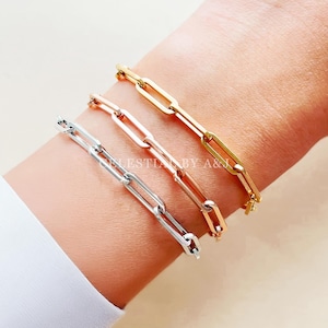 May include: Three gold, silver and rose gold chain bracelets with a box chain design. The bracelets are stacked on a person's wrist.