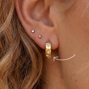 Könnte beinhalten: Nahaufnahme eines goldenen Creolen-Ohrrings mit Sternmuster und kleinen Ohrsteckern. Der Creolen-Ohrring wird am Ohr einer Person mit blondem Haar getragen. Die Ohrringe haben eine leuchtend goldene Farbe.