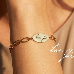 Może przedstawiać: Złota bransoletka z owalnym charms'em i ogniwami łańcucha. Charms ma ręcznie pisany tekst "Love Jim". Bransoletka jest noszona na nadgarstku.