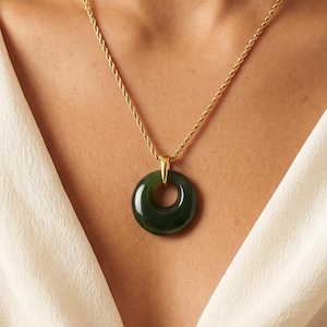 May include: A gold-toned rope chain necklace featuring a circular, dark green jade-like pendant. The pendant is suspended from a polished gold-toned metal bail, creating a simple and elegant piece of jewelry that rests against the neckline.