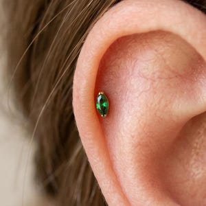 May include: A gold-toned metal earring featuring a marquise-cut green gemstone, secured in the upper cartilage of an ear. The jewelry piece has a slender, pointed oval shape with a polished metallic setting holding the vibrant green stone.