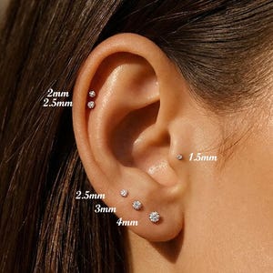 May include: Close-up of an ear with multiple small, sparkling stud earrings. The earrings range in size from 1.5mm to 4mm. The ear is set against a blurred background, highlighting the jewelry.