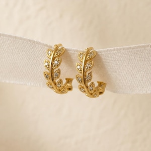 May include: A pair of gold-colored hoop earrings. Each earring features a leaf design with small, clear gemstones. The earrings are displayed on a white ribbon against a soft, neutral background.