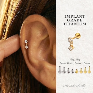 Może przedstawiać: Złoty kolczyk tytanowy z dwoma przezroczystymi, błyszczącymi kamieniami. Kolczyk jest pokazany na uchu, z tekstem "IMPLANT GRADE TITANIUM" i szczegółami rozmiaru: 5 mm, 6 mm, 8 mm, 10 mm. Sprzedawane pojedynczo.