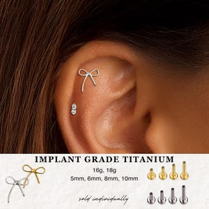 May include: A close-up of an ear with two pieces of jewelry. One is a silver bow-shaped earring, and the other is a small stud with three clear stones. The text "IMPLANT GRADE TITANIUM" is visible, along with size specifications.