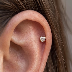 May include: A small gold stud earring featuring a clear, heart-shaped gemstone set in a four-prong setting. The earring is positioned in the upper cartilage area of an ear, showcasing a delicate and reflective design with a polished metal finish.