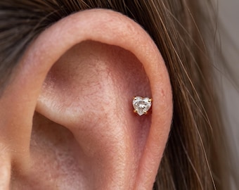 May include: A small gold stud earring featuring a clear, heart-shaped gemstone set in a four-prong setting. The earring is positioned in the upper cartilage area of an ear, showcasing a delicate and reflective design with a polished metal finish.