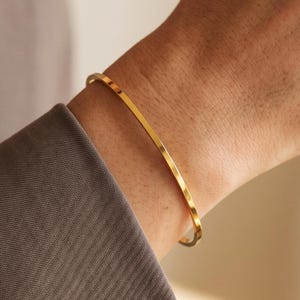 May include: A thin, polished gold-tone metal bangle bracelet with a smooth, flat surface and a circular shape. The bracelet features a simple, continuous band design with a reflective finish that catches the light, worn around the wrist.