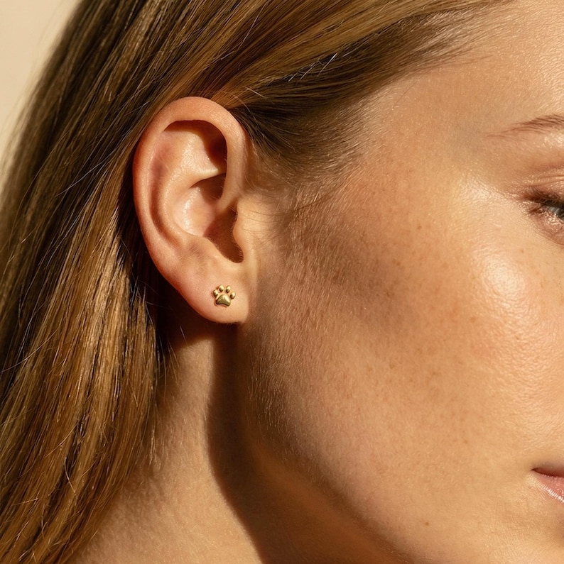 May include: A close-up of a person's ear with a small, gold-colored paw print stud earring. The earring is a detailed representation of a paw print. The person has light skin and long, light brown hair.