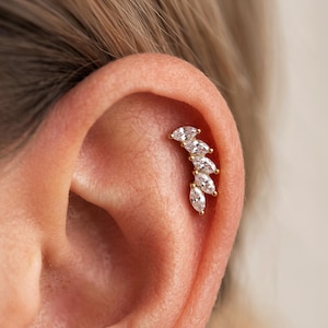 May include: A gold-toned ear cartilage earring featuring five marquise-cut clear cubic zirconia stones arranged in a curved, ascending line. The stones are set in a metal frame that follows the natural contour of the upper ear cartilage.