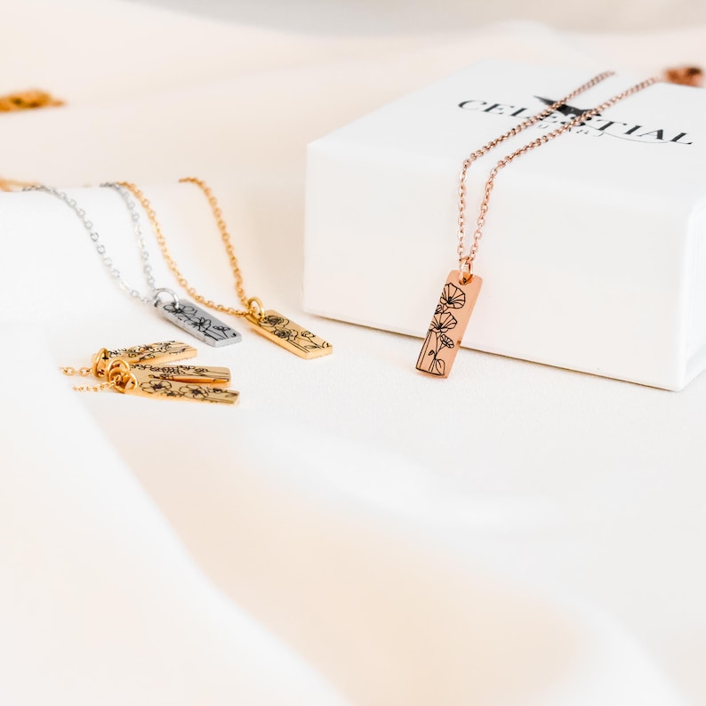 May include: Three gold, silver, and rose gold necklaces with delicate chains and rectangular pendants featuring a floral design. The necklaces are displayed on a white surface with a white box that says "Celestial Art".