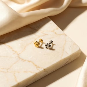 May include: Two paw-shaped stud earrings. One is gold-colored, the other is silver-colored. The earrings are displayed on a marble surface with a soft, cream-colored fabric in the background. The earrings are small and delicate.
