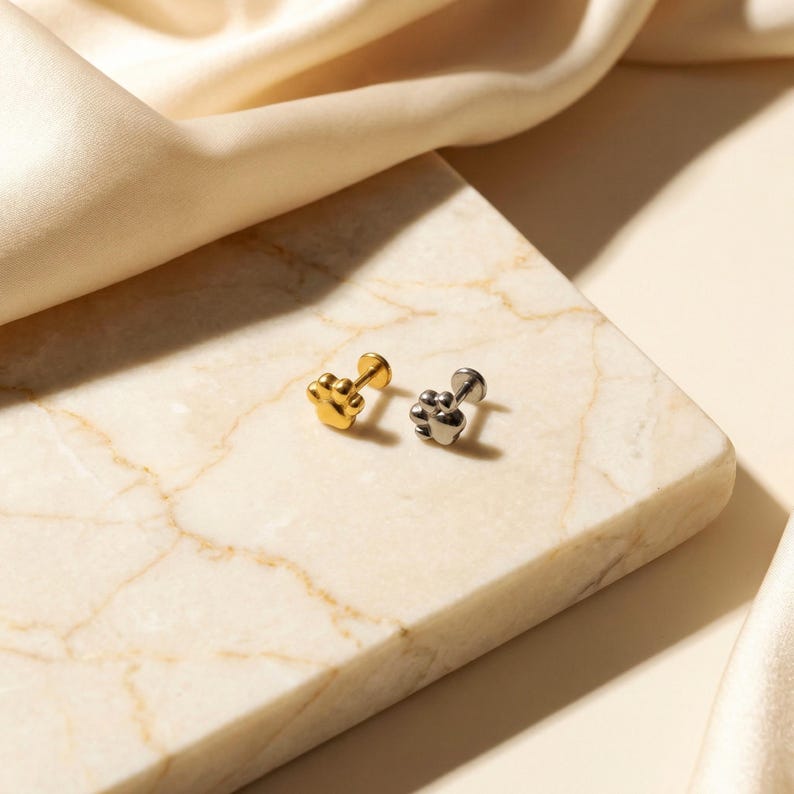 May include: Two paw-shaped stud earrings. One is gold-colored, the other is silver-colored. The earrings are displayed on a marble surface with a soft, cream-colored fabric in the background. The earrings are small and delicate.
