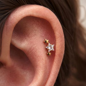 May include: A gold-tone ear cartilage stud featuring a cluster of three stars. The central star is larger and set with a clear, faceted cubic zirconia stone, while the two smaller stars above and below are solid gold-tone metal.
