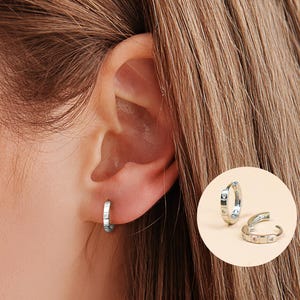 May include: Silver hoop earrings with small square and rectangular accents. One earring is worn on the ear, and two additional earrings are displayed in a circular inset. The earrings have a simple, elegant design.