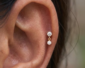 May include: A gold earring featuring two small, round, clear faceted stones set in a vertical drop design. The top stone is secured to the ear, while the second stone hangs below it, connected by a small gold link, creating a delicate, dangling appearance.