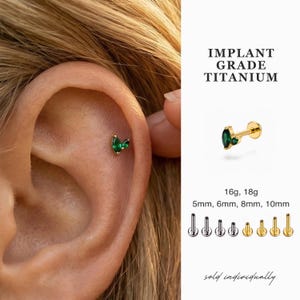 Puede incluir: Un pendiente de titanio dorado de grado implante con una gema verde esmeralda con corte marquesa. La imagen muestra el pendiente en uso y una foto del producto. El texto dice "IMPLANT GRADE TITANIUM" y "sold individually."