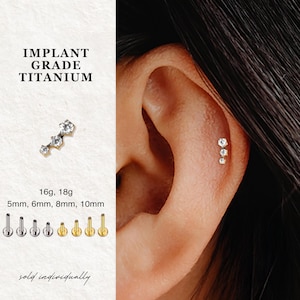 May include: A close-up of an ear with three small, sparkling stud earrings. The earrings are set with clear gemstones. The image also shows a separate earring with three gemstones and text that reads "IMPLANT GRADE TITANIUM", "16g, 18g", "5mm, 6mm, 8mm, 10mm", and "sold individually".