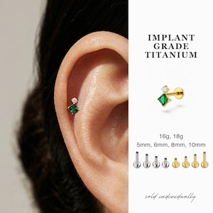 May include: A gold-colored titanium earring with a small, square, emerald-green gemstone and a small, round, clear gemstone. The earring is shown on an ear. Text on the image reads "IMPLANT GRADE TITANIUM".