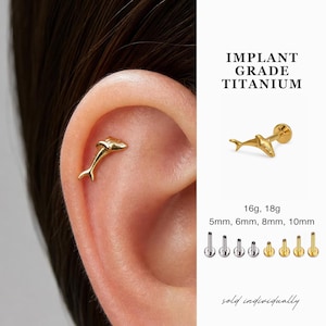 May include: Gold-coloured dolphin-shaped ear piercing stud. The image shows the stud in an ear and a separate image of the stud. Text on the image reads "IMPLANT GRADE TITANIUM" and "16g, 18g" with sizes 5mm, 6mm, 8mm, and 10mm.