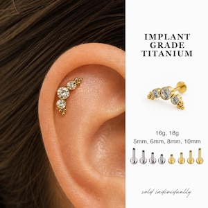 May include: Gold-colored titanium ear piercing jewelry with clear gemstones. The jewelry is shown on an ear and also displayed separately. The text reads "IMPLANT GRADE TITANIUM" and lists sizes 16g, 18g, 5mm, 6mm, 8mm, and 10mm.