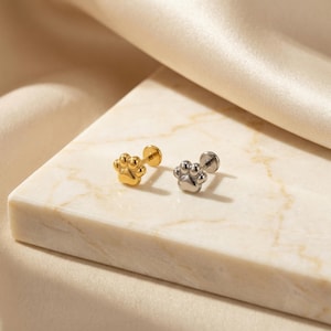 May include: Two paw-shaped stud earrings, one gold-colored and one silver-colored, are displayed on a marble surface. The earrings have a flat back and a post for piercing. The background is a soft, neutral color.