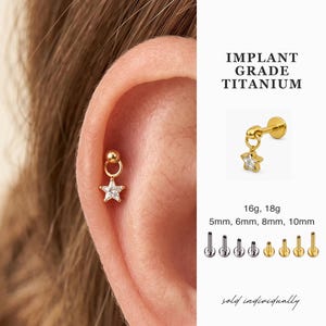 Puede incluir: Un pendiente de titanio dorado con un dije en forma de estrella, que se muestra en una oreja. El pendiente tiene un pequeño perno de bola y una estrella colgante con una piedra transparente. El texto dice "IMPLANT GRADE TITANIUM".