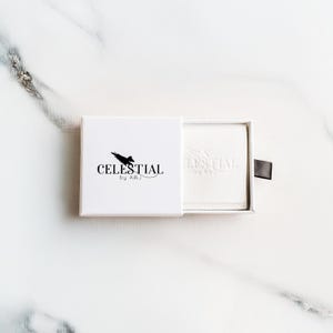 May include: A white box with the text "CELESTIAL by ABJ" in black. The box is partially open, revealing a white product inside. A black pull tab is attached to the box. The background is a white marble surface.