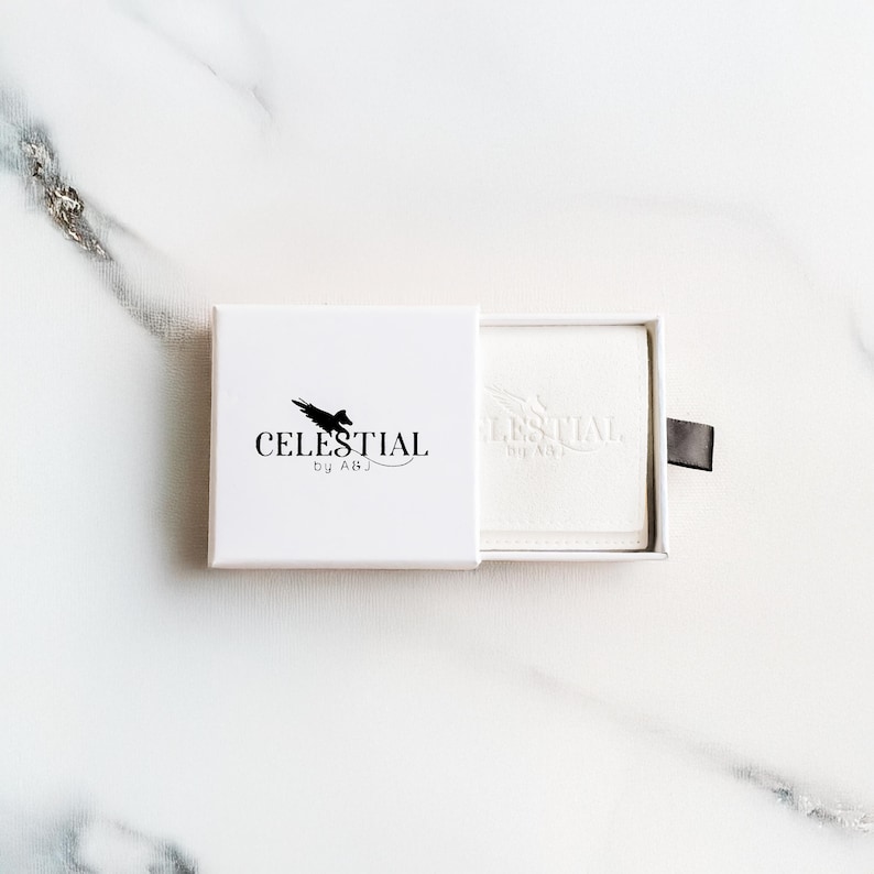 May include: A white box with the text "CELESTIAL by ABJ" in black. The box is partially open, revealing a white product inside. A black pull tab is attached to the box. The background is a white marble surface.