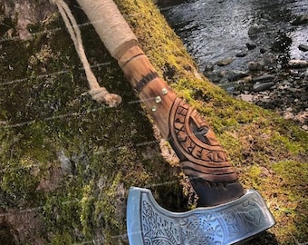 May include: A hand-carved wooden axe handle with intricate details and brass accents. The axe head is made of steel and features a detailed design with a Viking-inspired motif.