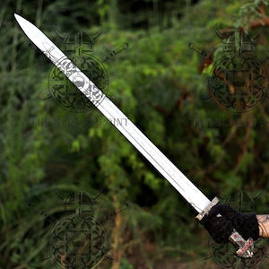 May include: A long, silver-toned metal sword featuring an intricate etched design on the blade near the hilt. The handle is detailed with metallic accents and small red circular inlays, held by a hand wearing a black textured glove.
