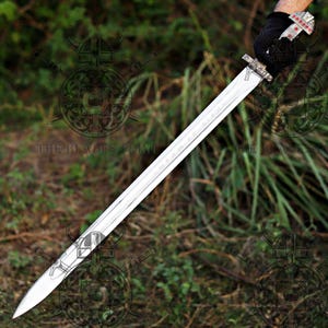 May include: A long, double-edged steel sword with a pointed tip and a central fuller. The hilt features a silver-toned crossguard and a pommel decorated with small red circular inlays. The blade is held by a hand wearing a black glove.