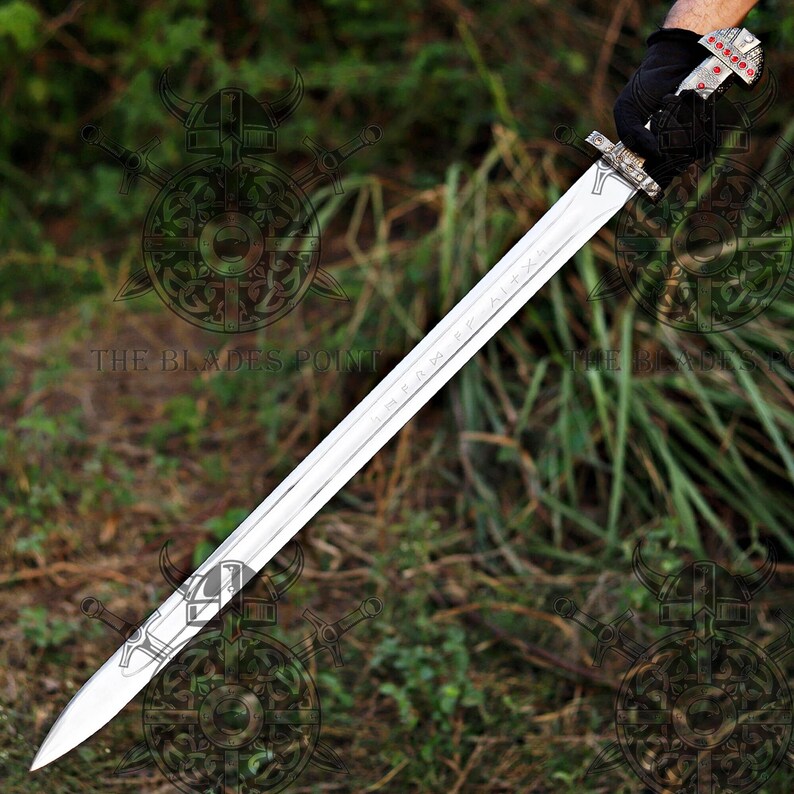 May include: A long, double-edged steel sword with a pointed tip and a central fuller. The hilt features a silver-toned crossguard and a pommel decorated with small red circular inlays. The blade is held by a hand wearing a black glove.